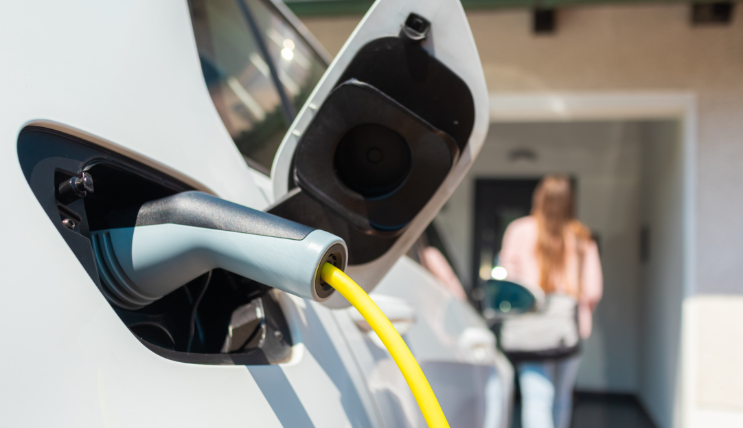 Ein Ladestecker steckt in einem E-Auto, im Hintergrund geht eine Frau zur Haustür – steht für komfortables und nachhaltiges Laden des Elektroautos mit Ökostrom direkt zuhause.
